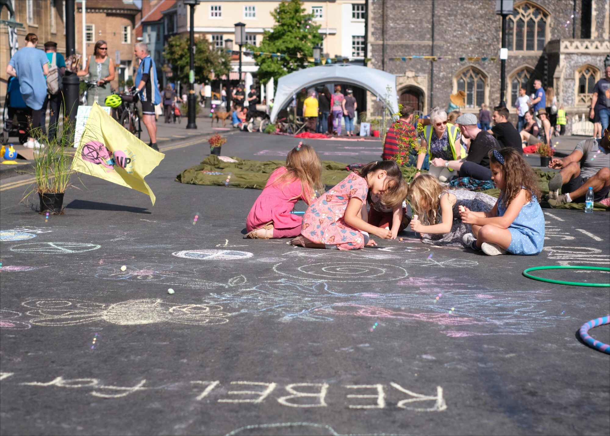 Children play and sit on a street on a sunny day. They have drawn in chalk on the tarmac.