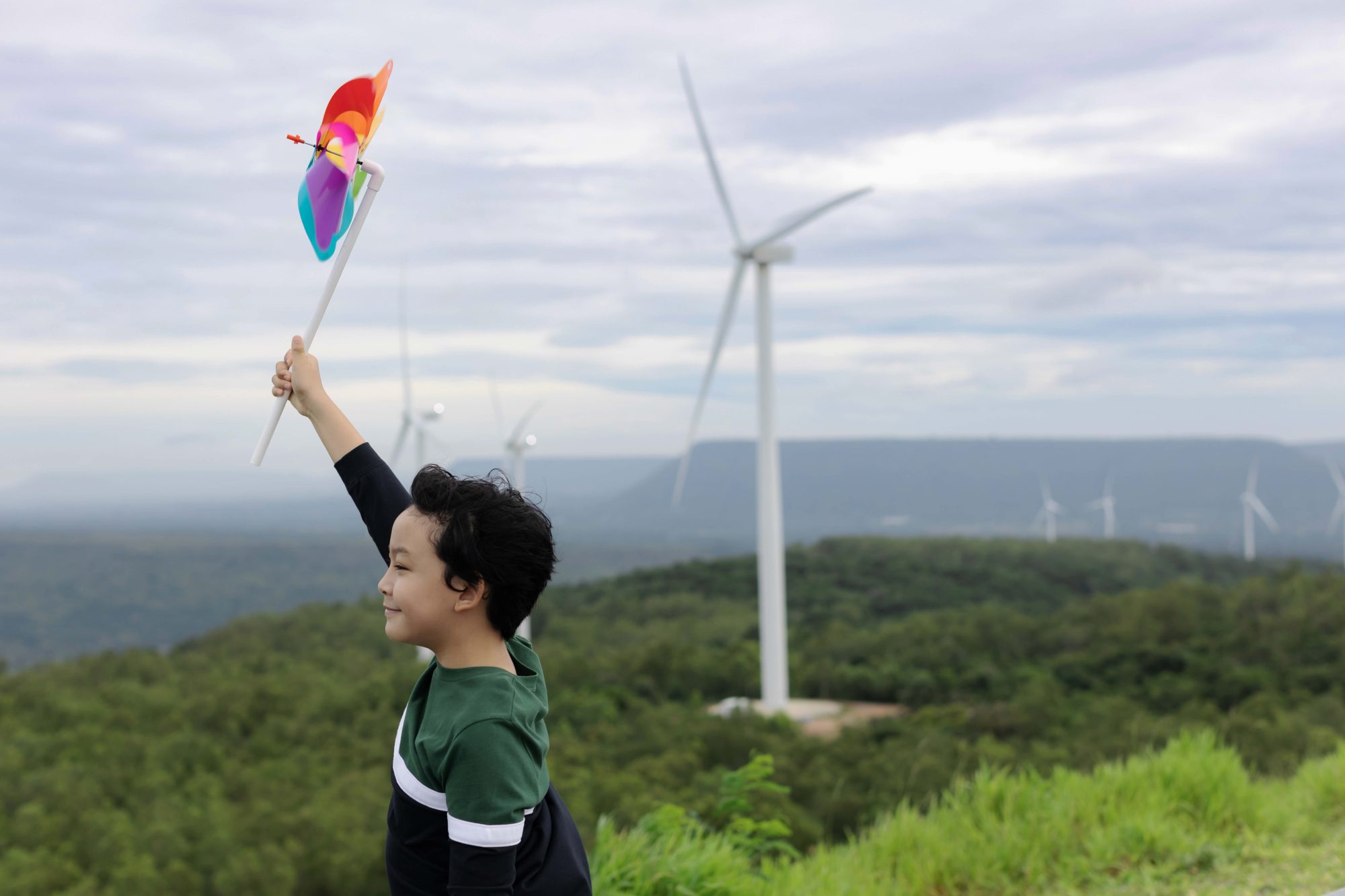 Young boy playing with wind pinwheel toy in the wind turbine farm, green field over the hill.