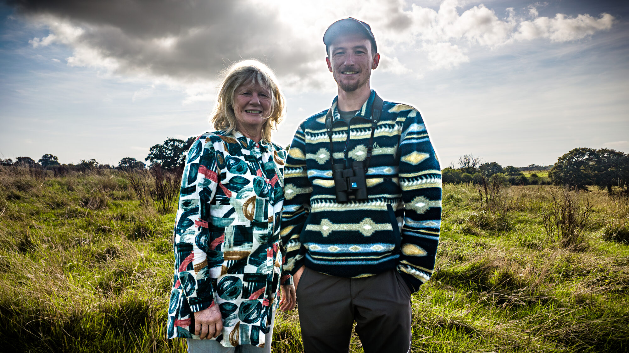 Jan Stannard co-founder at Heal Rewilding, and Dan Hill, Rewilding Ranger, stand on-site at Heal Somerset. Behind them is long rough grass and other meadow plants and a blue sunny sky with a white cloud.