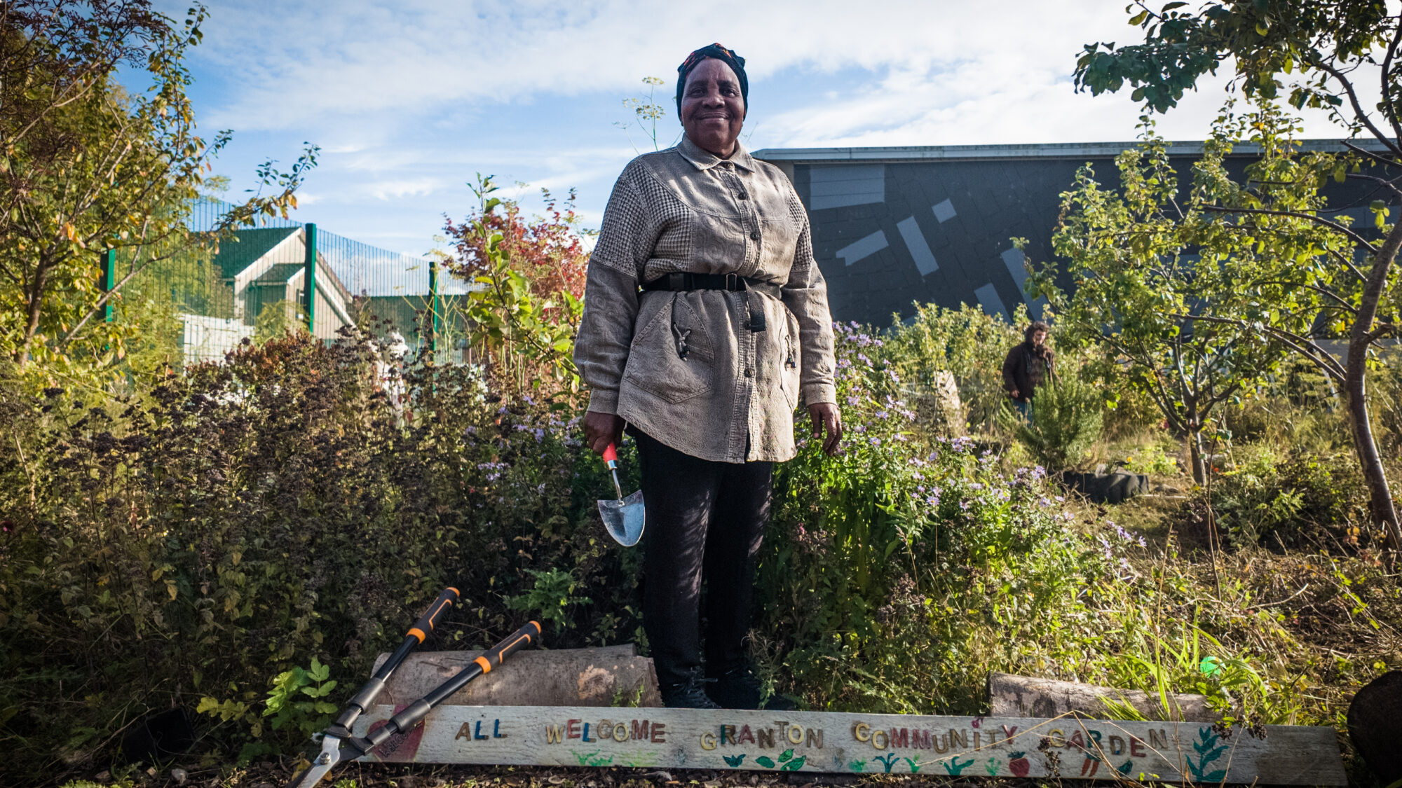 Mary, cofounder of Granton Community Gardeners, one of the projects being supported by Nature Neighbourhoods. Mary, an older lady originally from Kenya, is standing in the garden holding a trowel. It is a sunny day and she is surrounded by greenery. Below her is a sign saying "All Welcome Granton Community Garden"