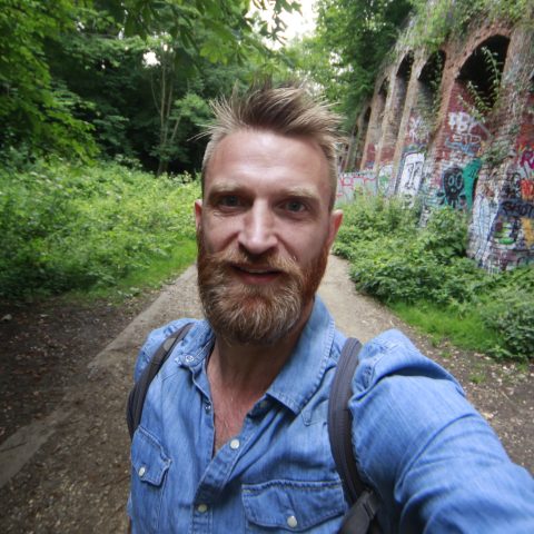 Daniel Raven-Ellison, founder of active travel network Slow Ways. A portrait photo of a man with blonde hair and a beard wearing a blue denim shirt. Behind him is a path with greenery growing on either side and a red brick viaduct on the right.