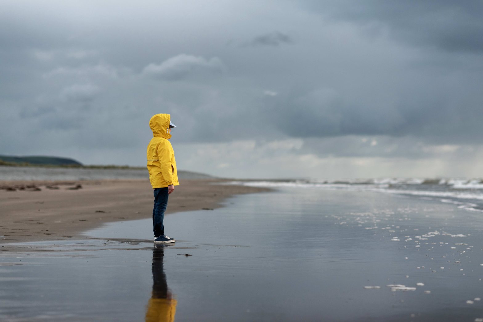 Boy in yellow jacket standing by stormy sea, looking at sea.