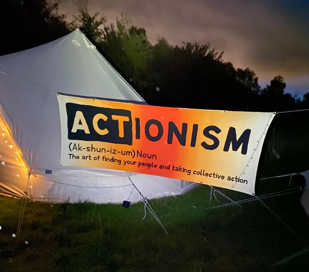 A white tent in a dark field with a brighly coloured banner tied to it reading: Actionism - (Ak-shun-iz-um) Noun The art of finding your people and taking collective action.