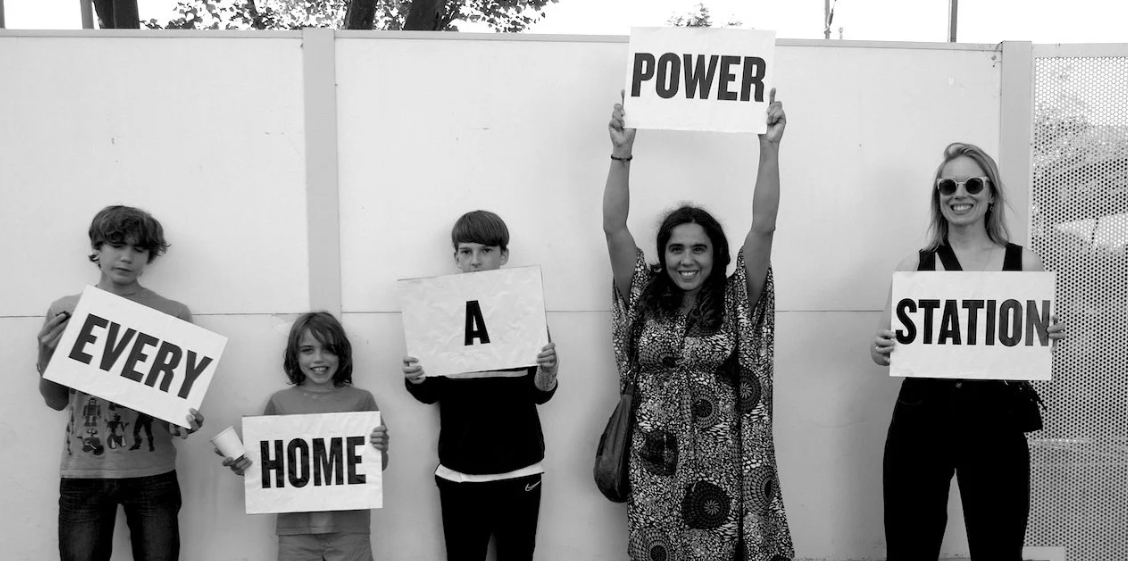 Smiling participants of "Power Station" - an artistic project about bringing locally generated electricity to households along a street in London. The people in the photo hold signs which together say "Every Home A Power Station".