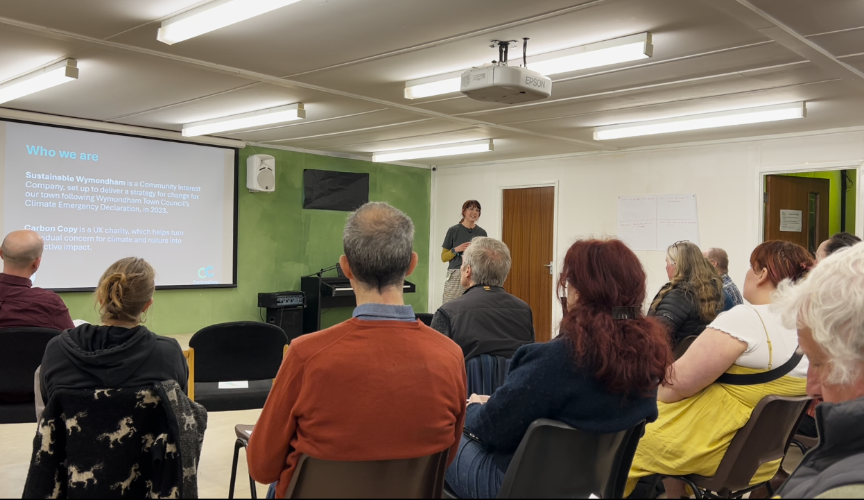 Isabelle Sparrow stands in front of an audience at Rothbury Community Hall at the Wymondham People's Emergency Briefing event. Behind her is a screen with text projected onto it with information about Sustainable Wymondham and Carbon Copy.