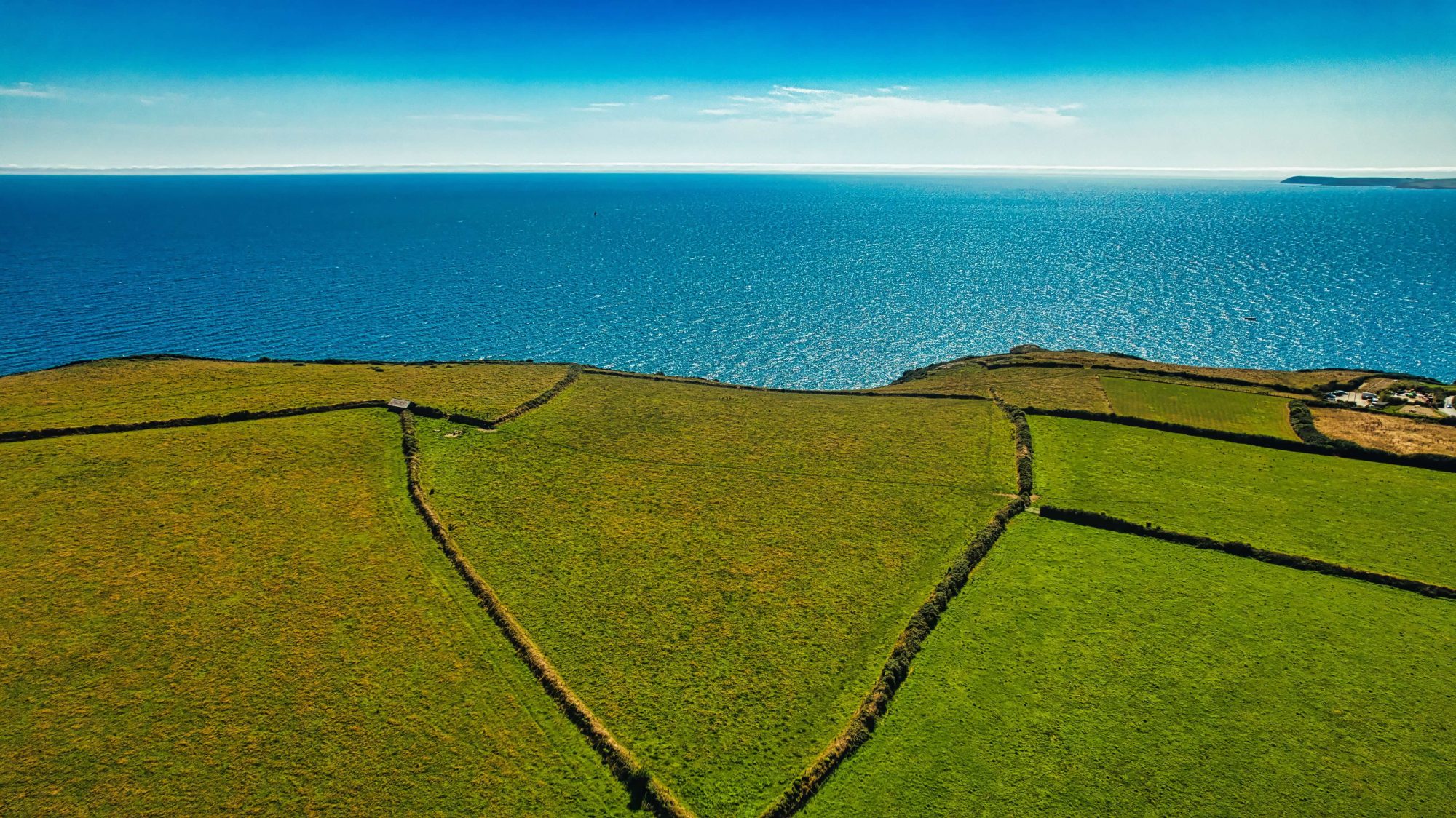 Aerial view of green fields meeting the sea. The landscape features distinct geometric patterns of farmland bordered by hedgerows, with the horizon blending into the sea.